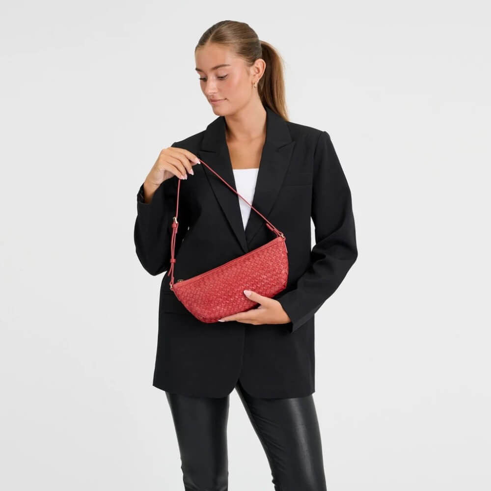 A woman wearing a black blazer and black pants holds a Depeche red woven-texture shoulder bag with a thin red strap against a plain light grey studio background.