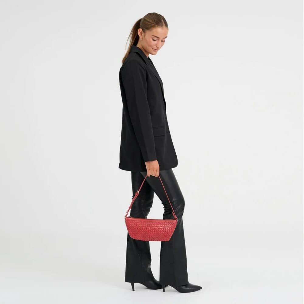 A woman in a black blazer, black pants, and black heels holds a Depeche red woven leather shoulder bag against a plain white studio background.