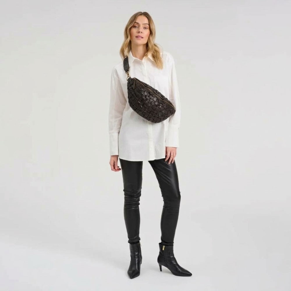 A woman modelling a Depeche dark brown woven crossbody fanny pack worn over her shoulder, styled with a white button-down shirt, black leather pants, and black ankle boots against a light grey studio background.
