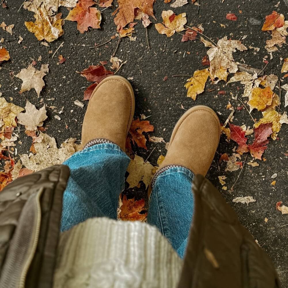 Person wearing brown EMU and blue jeans standing on a ground covered with autumn leaves. EMU Sharky Scuff Chestnut