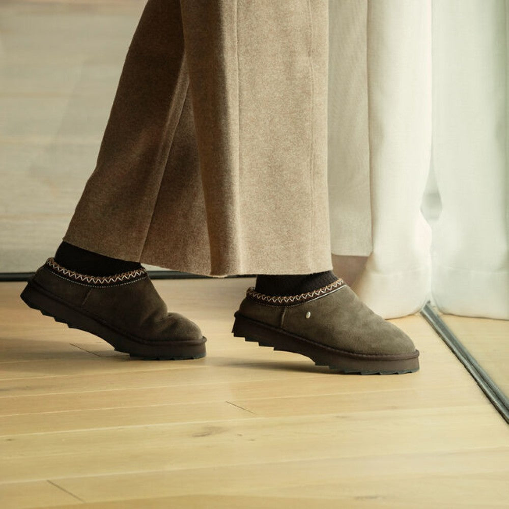 A person wearing Emu dark brown suede Sheepskin Slippers, paired with black socks and wide-leg taupe trousers, standing on a light wood floor near a white curtain.
