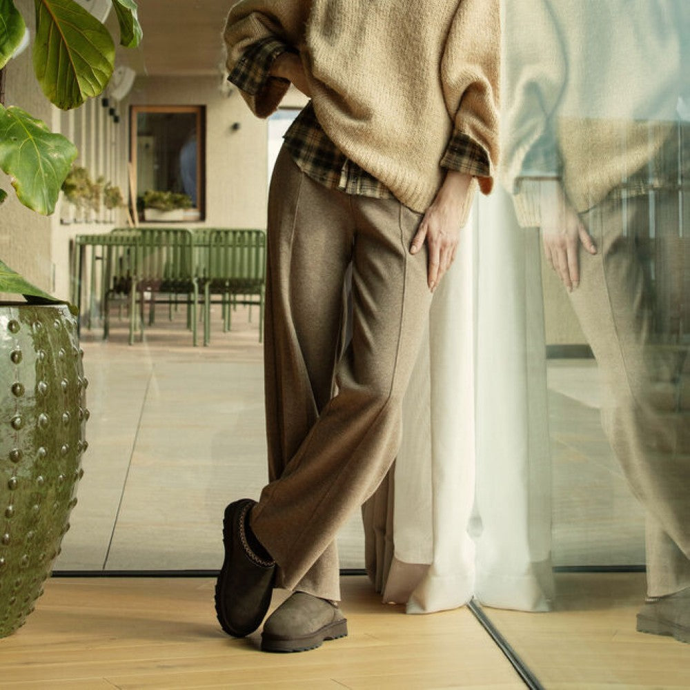 A person wearing a beige knit sweater, brown wide-leg trousers, and Emu dark slip-on slippers stands indoors near a glass wall with a mirror reflection. The setting includes a large green potted plant and a modern interior background.