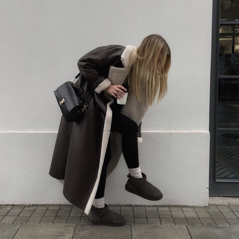 Person wearing a long dark coat with white fur trim, black pants, and brown shoes, leaning against a light gray wall. EMU Stinger Micro Chocolate