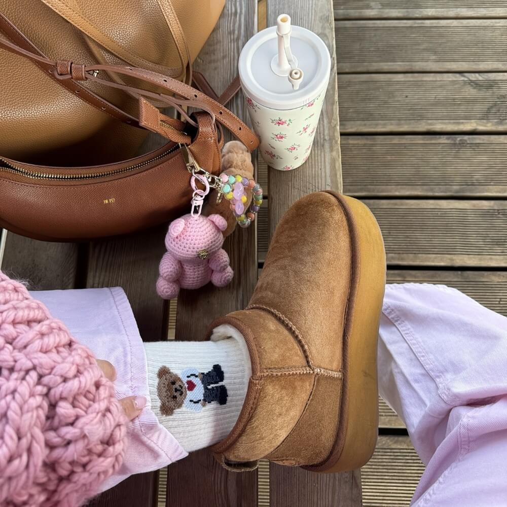 Brown slip-on shoe with white fur lining, pink knit sleeve, and a small teddy bear keychain on a wooden surface. EMU Stinger Micro Flatform Chestnut