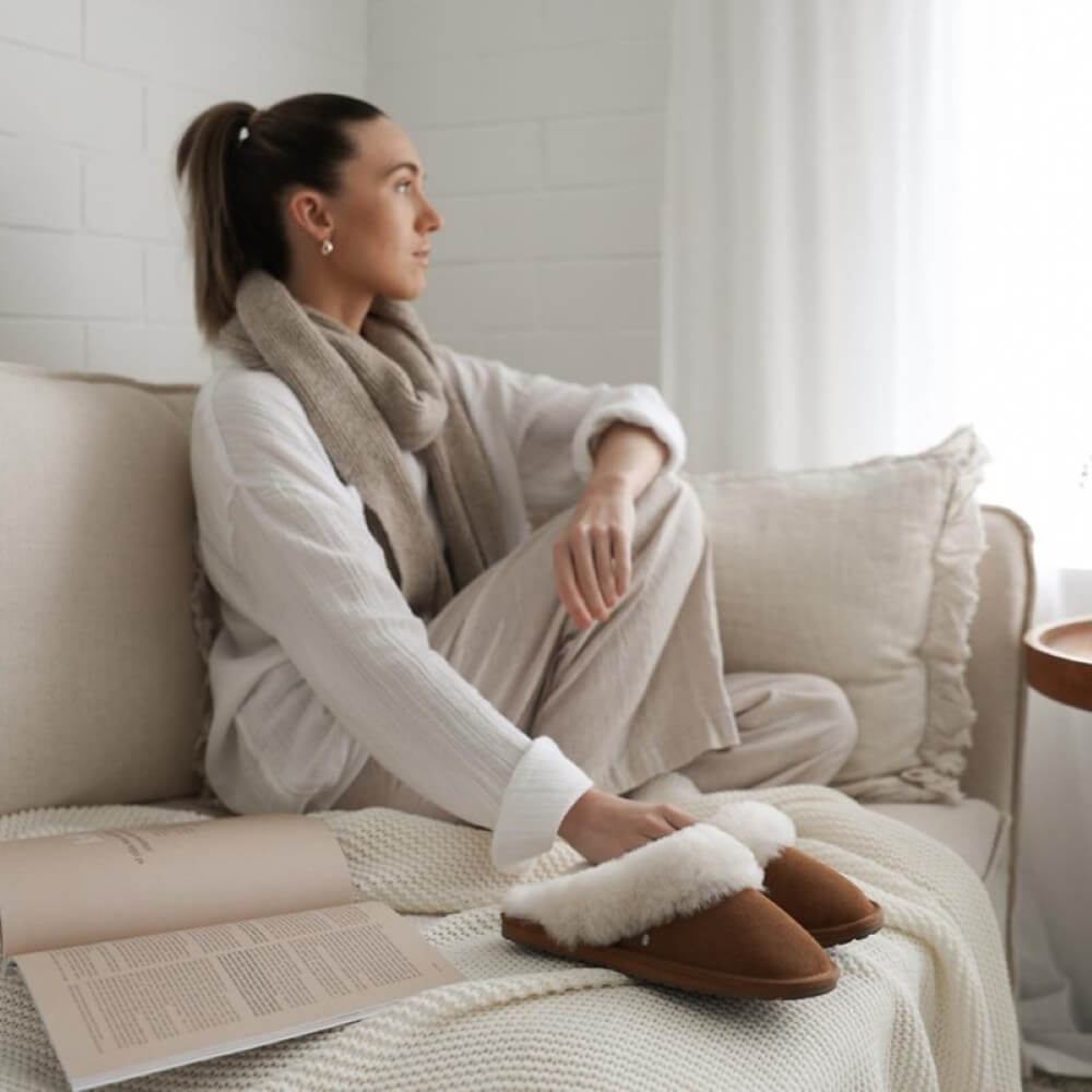 Woman sitting on a couch wearing slippers and a scarf, with an open book on her lap. EMU Australia Jolie Slipper Chestnut