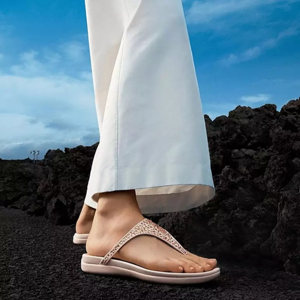 Person wearing beige fitflop sandals on a rocky surface with a blue sky.