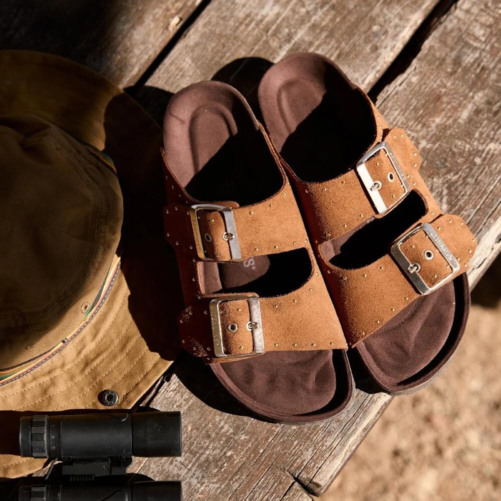Genuins Brown leather sandals with buckles on a wooden surface next to a brown jacket and black binoculars.