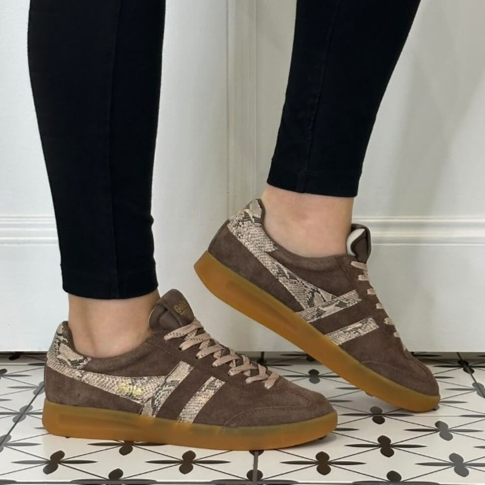 A person wearing Gola brown suede sneakers with snakeskin-patterned side stripes and gum soles, standing on a black-and-white patterned tile floor against a white wall.