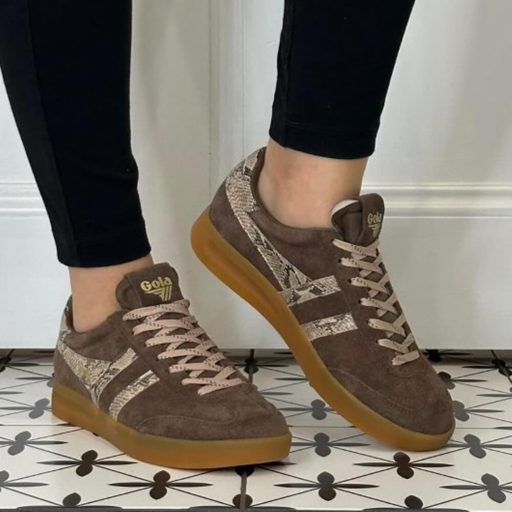 A person wearing Gola brown suede sneakers with snakeskin-patterned side stripes and gum soles, standing on a black-and-white patterned tile floor against a white wall.