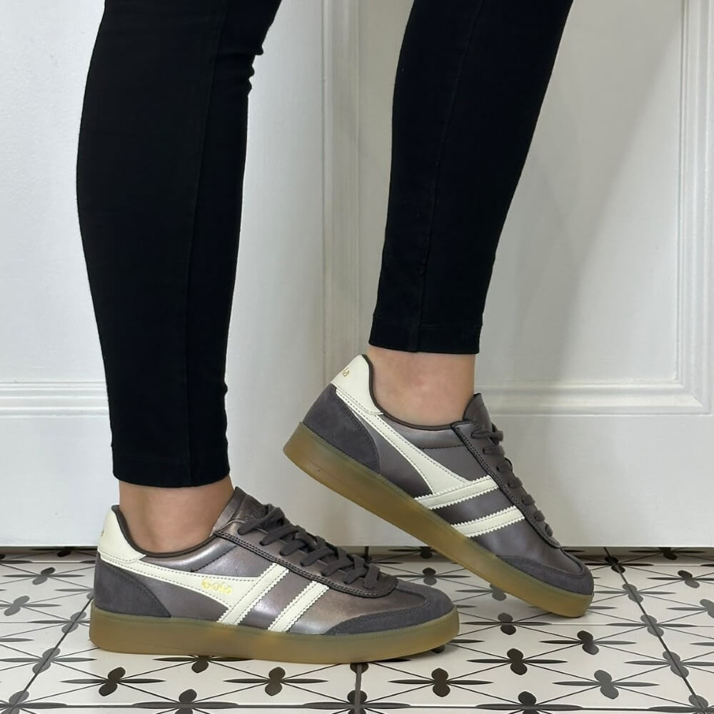 A person wearing black leggings and Gola grey low-top sneakers with white side stripes and gum soles, standing on a patterned tile floor in front of a white panelled wall.