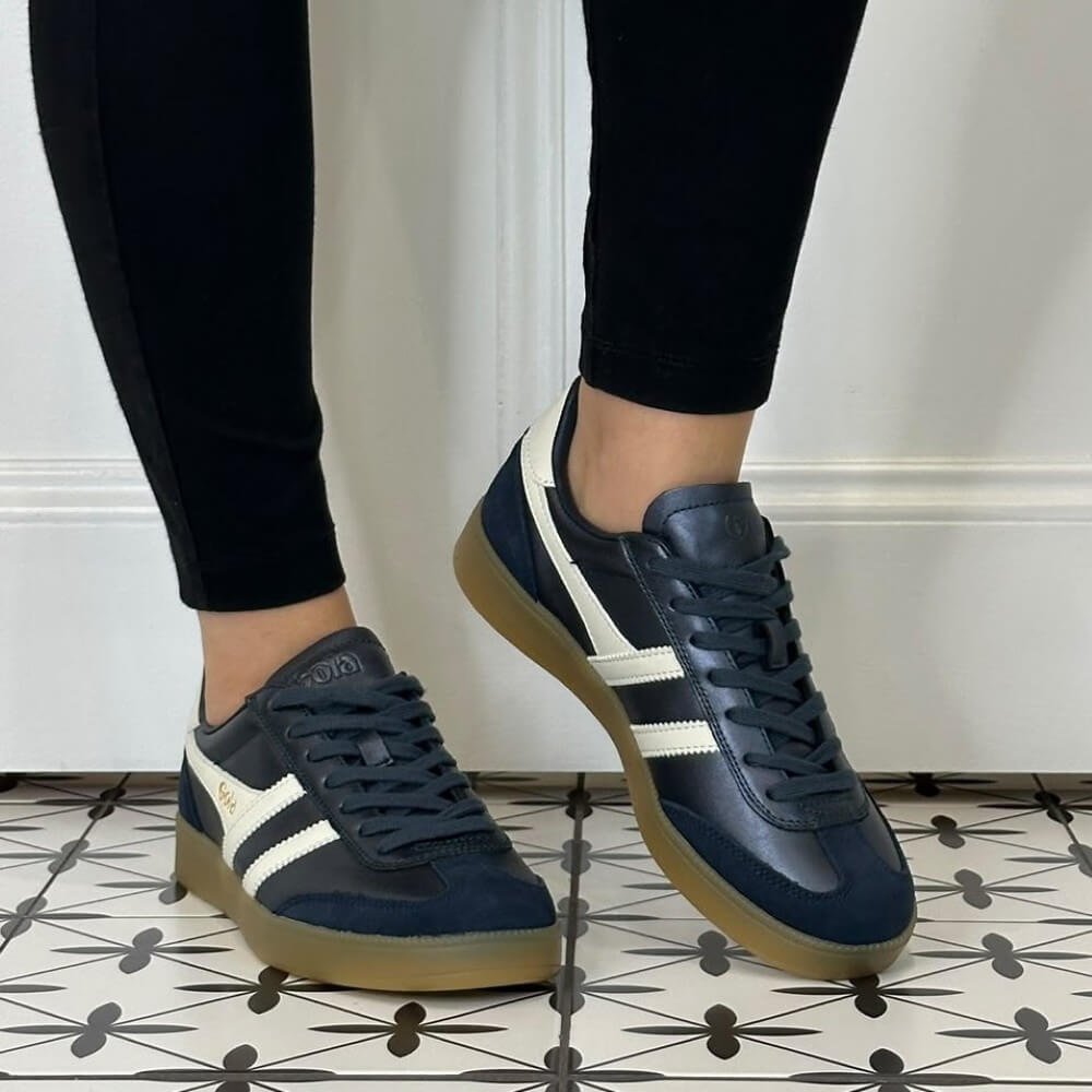 A person wearing Gola navy blue and off-white sneakers with gum soles, paired with black leggings, standing on a patterned tile floor in front of a white panelled wall.