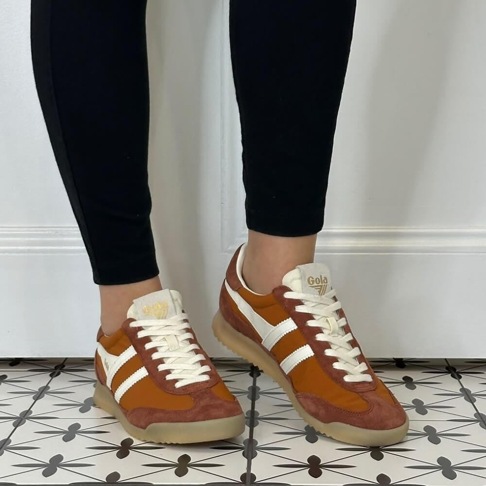 A person wearing Gola brown suede and canvas low-top sneakers with white stripes and white laces, paired with black leggings, standing on a black and white patterned tile floor against a white wall.