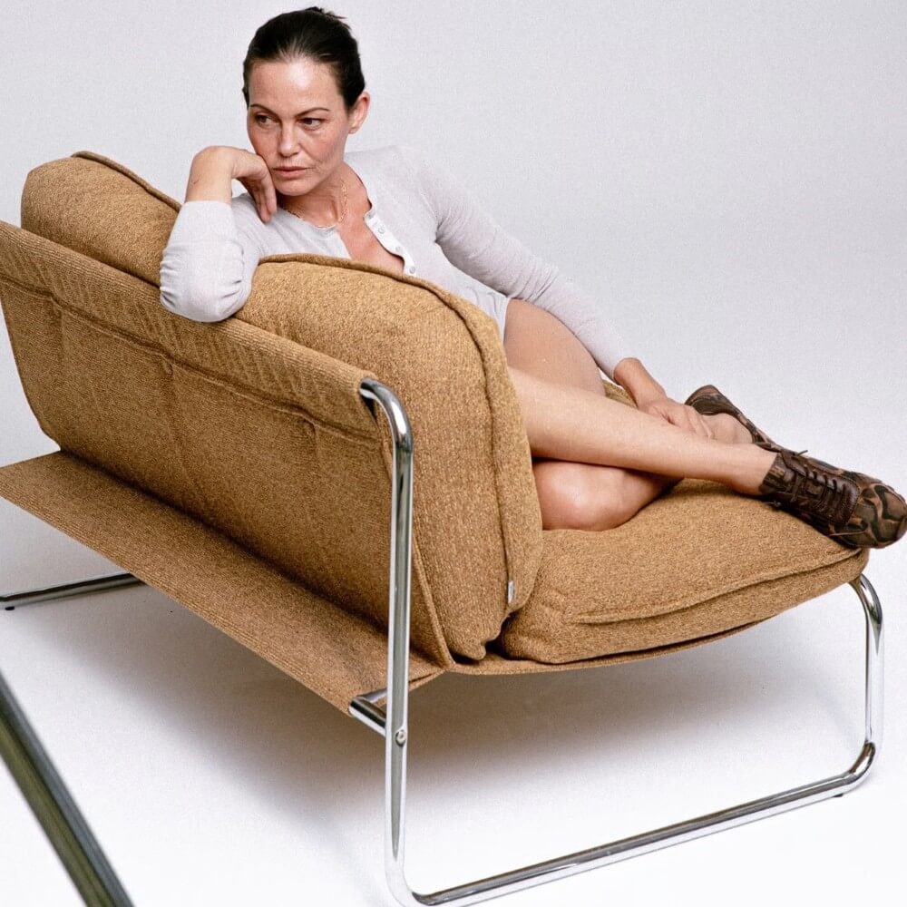 A woman reclining on a modern tan upholstered lounge chair Hispanitas Trainers against a plain light grey background.