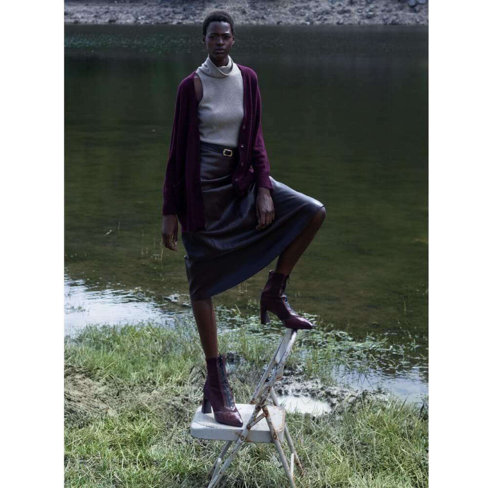 Woman standing on a small white stool by a body of water wearing a purple cardigan and skirt.