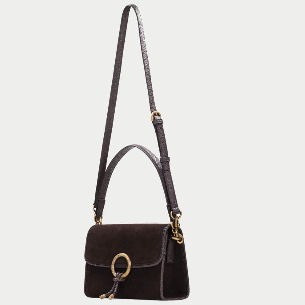 A Hispanitas dark brown suede shoulder bag with a gold-tone circular ring detail and a matching leather tassel, shown against a light gray
