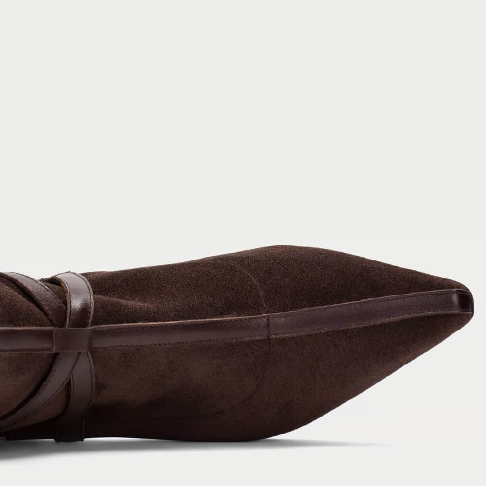 Close-up side view of a dark brown suede pointed-toe boot with a thin dark brown leather band detail, set against a plain light gray background.
