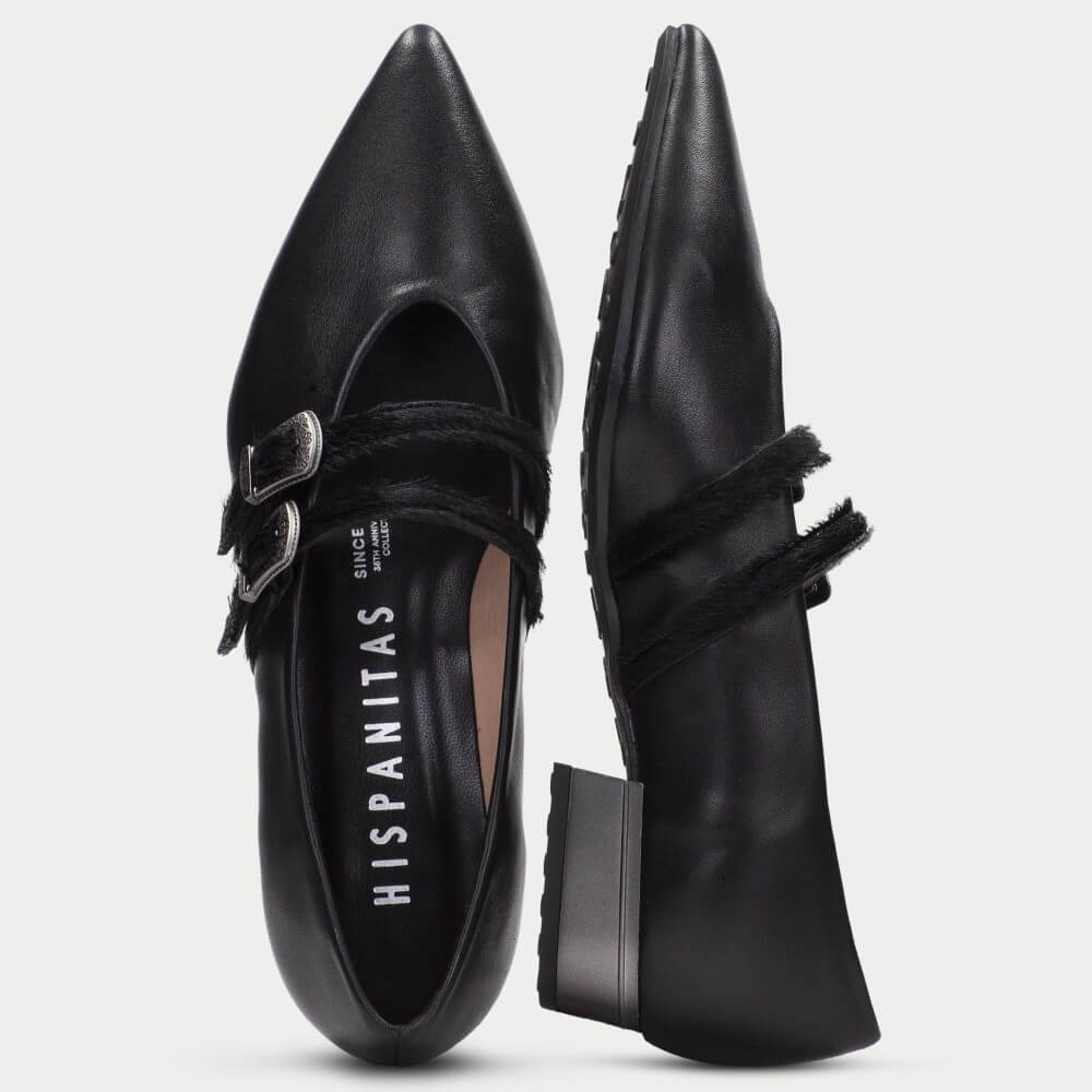 A pair of black leather pointed-toe flat shoes by Hispanitas, shown from the side and back. One shoe features a double strap design with a silver buckle, and the other shows the side profile with a black fringe detail across the instep.