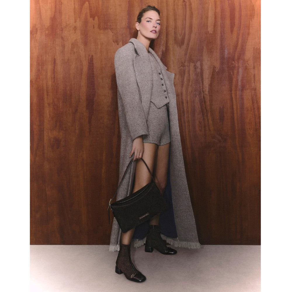 A model stands against a wooden wall wearing a long gray coat over a matching gray vest and shorts, holding a black handbag.