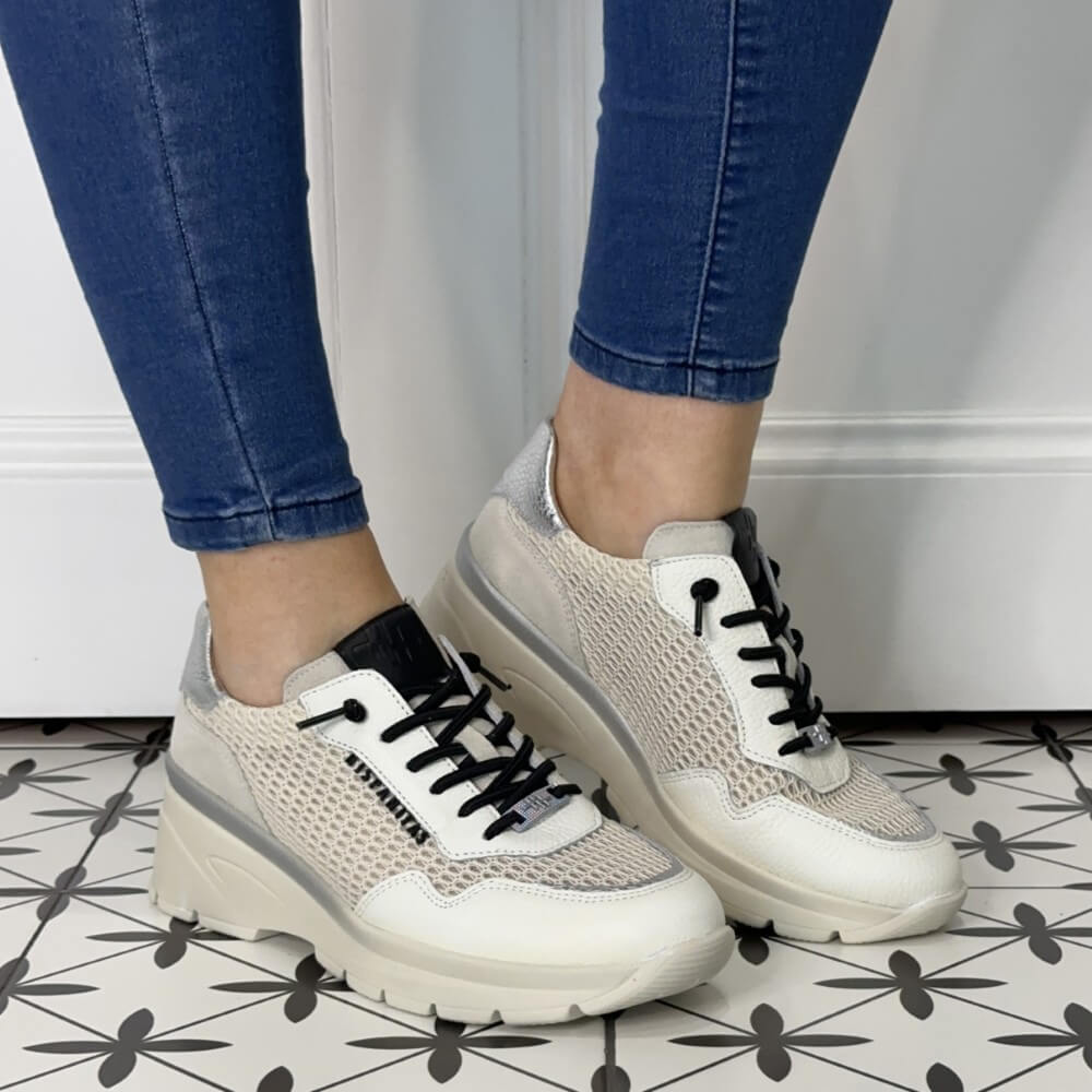 Beige sneakers with black laces worn with blue jeans on a tiled floor.