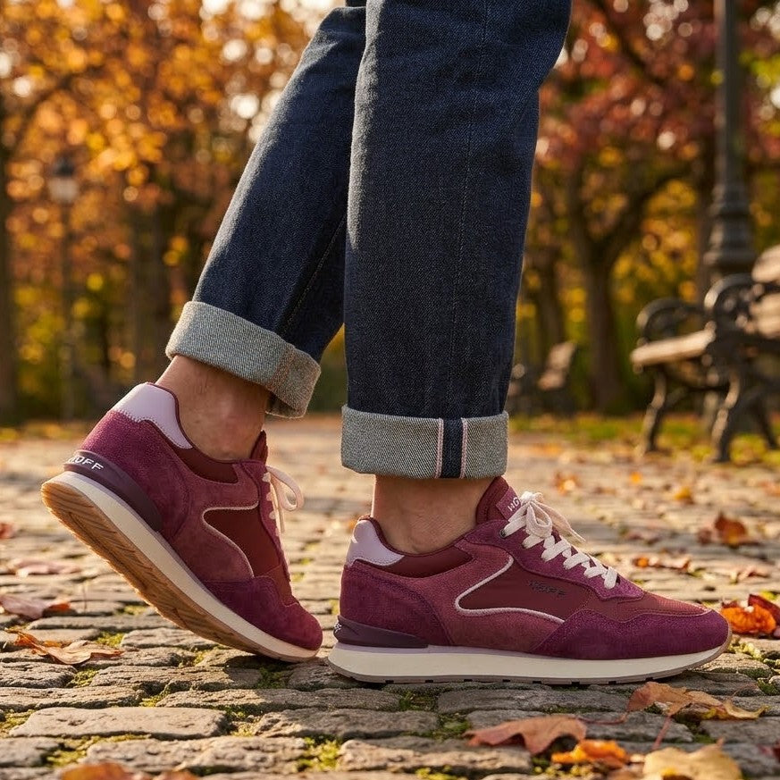 A person wearing cuffed blue jeans and Hoff burgundy suede sneakers with white laces, standing on a cobblestone path with autumn leaves and trees in the background.