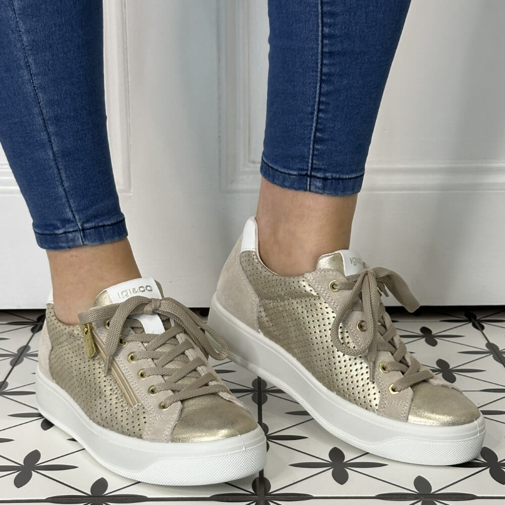 Gold sneakers with white soles worn with blue jeans on a patterned floor.
