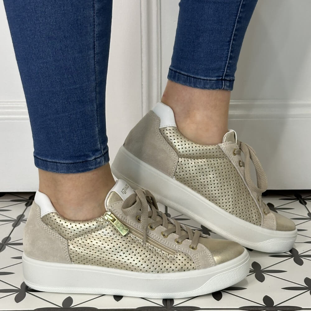 Gold sneakers with white soles worn with blue jeans on a patterned floor.