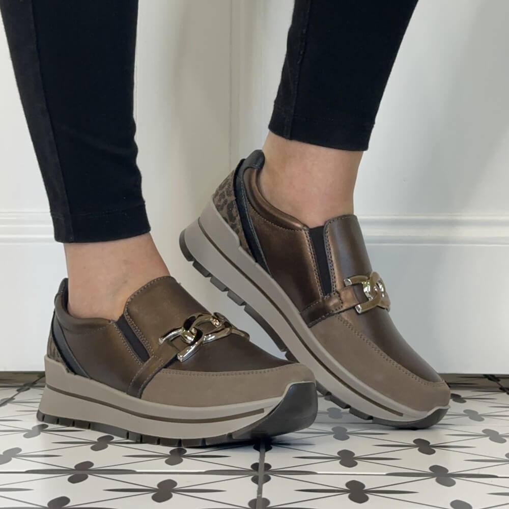 A person wearing Igi & Co Taupe slip-on platform sneakers with a decorative buckle detail, paired with black pants, standing on a patterned tile floor.