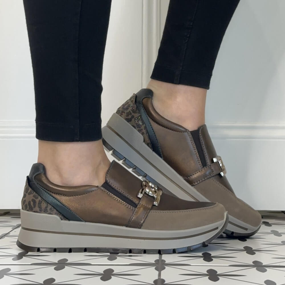 A person wearing Igi & Co Taupe slip-on platform sneakers with a decorative buckle detail, paired with black pants, standing on a patterned tile floor.