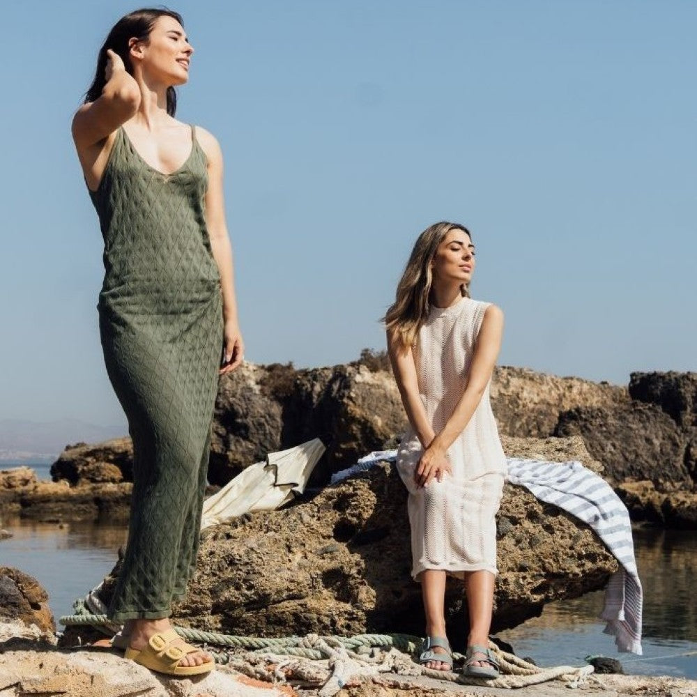 Two women standing on rocky terrain by a body of water, wearing sleeveless dresses.