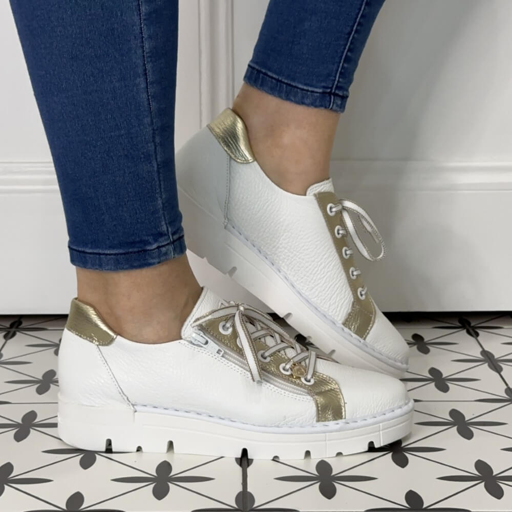 White sneakers with gold accents worn by a person on a patterned floor.