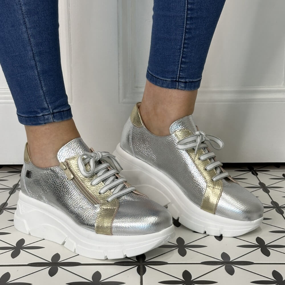 Silver sneakers with gold accents worn by a person on a patterned floor.