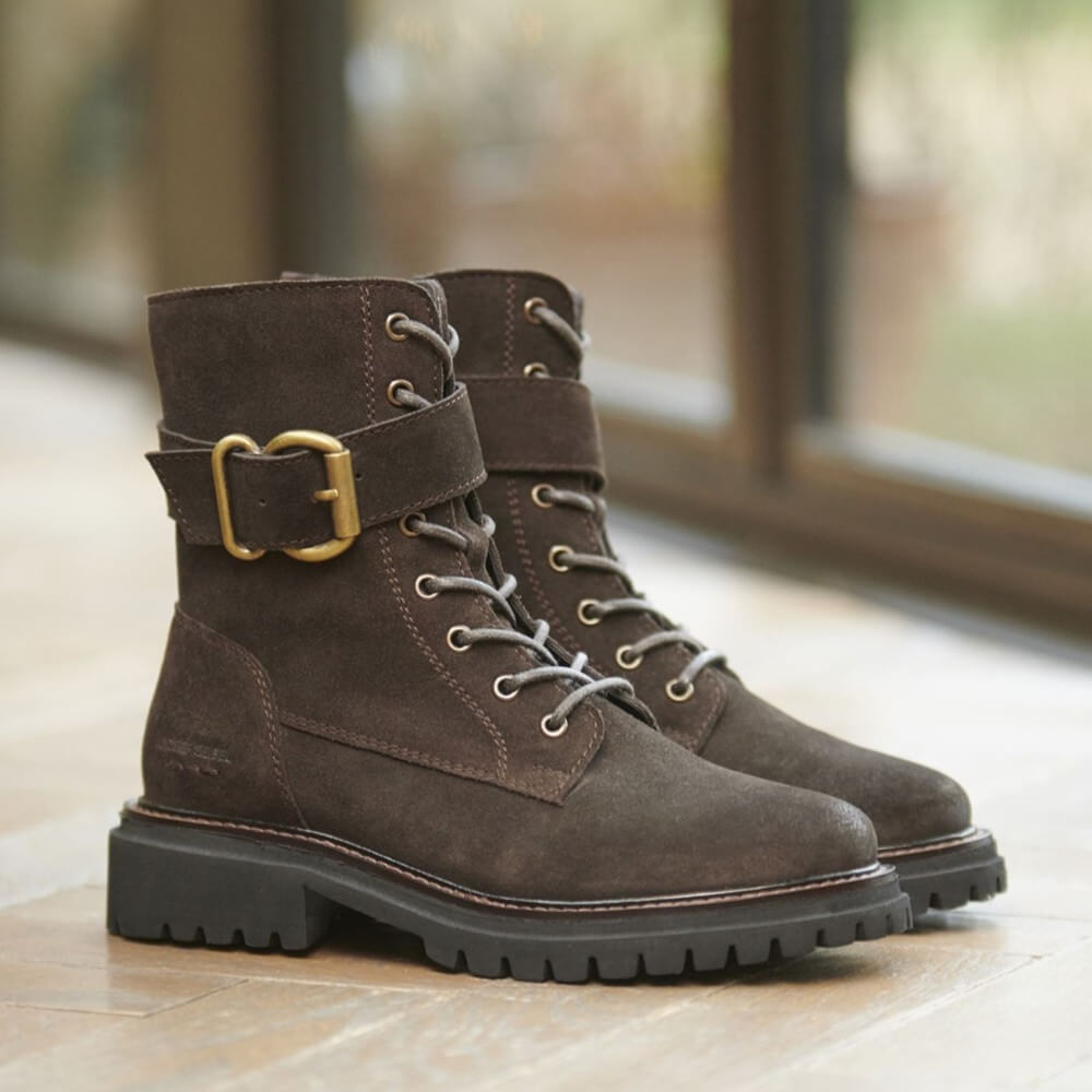 A pair of Josef Seibel dark brown suede lace-up boots with a wide strap and gold buckle on the front boot, resting on a wooden floor in front of a softly blurred window.