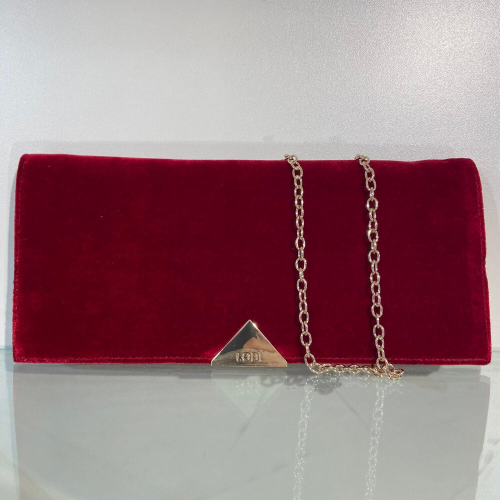 A Lodi red velvet clutch bag with a gold chain strap and a triangular gold metal accent on the front, displayed on a reflective white surface.