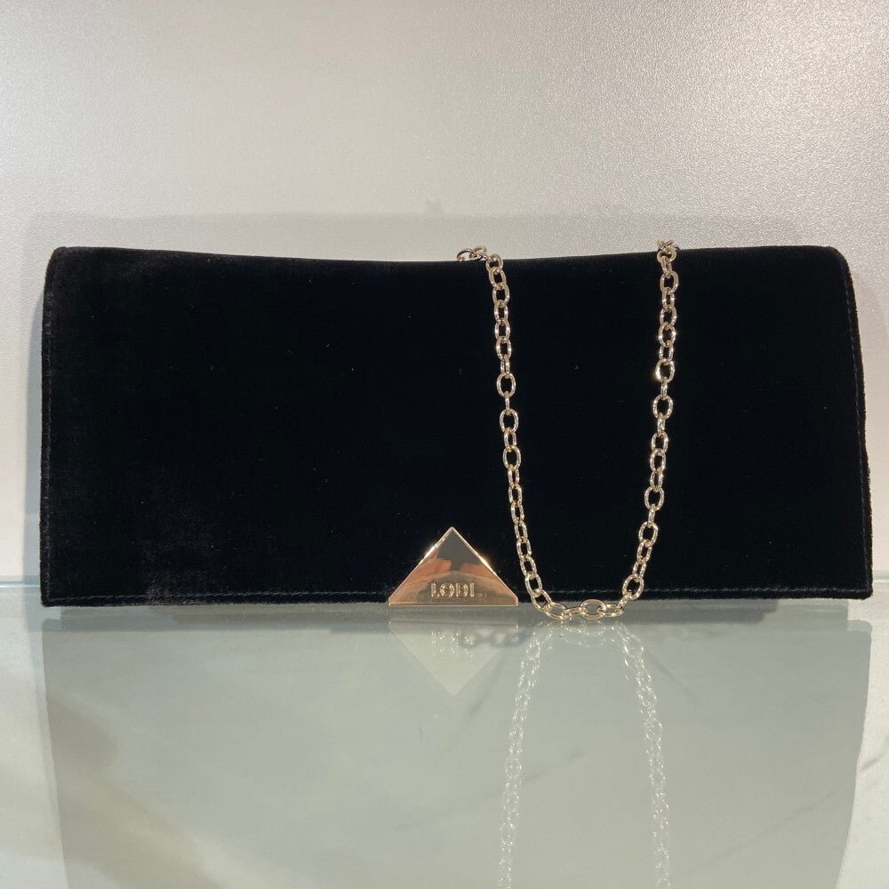 A Lodi black velvet clutch bag with a gold-tone chain strap and a triangular gold-tone metal emblem on the front, displayed on a reflective surface against a light background.