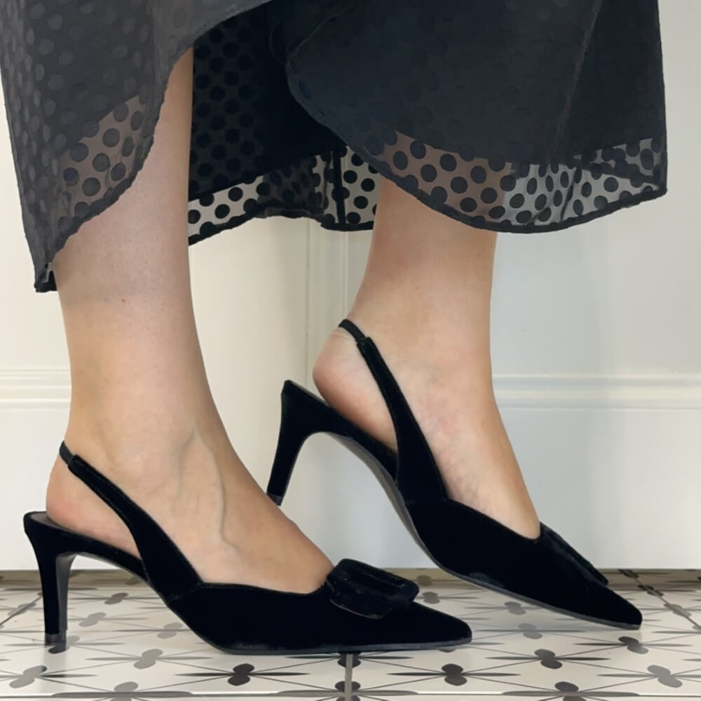 A person wearing Lodi black Velvet pointed slingback kitten-heel pumps with a small bow detail, paired with a sheer black polka-dot dress, standing on a patterned tile floor.