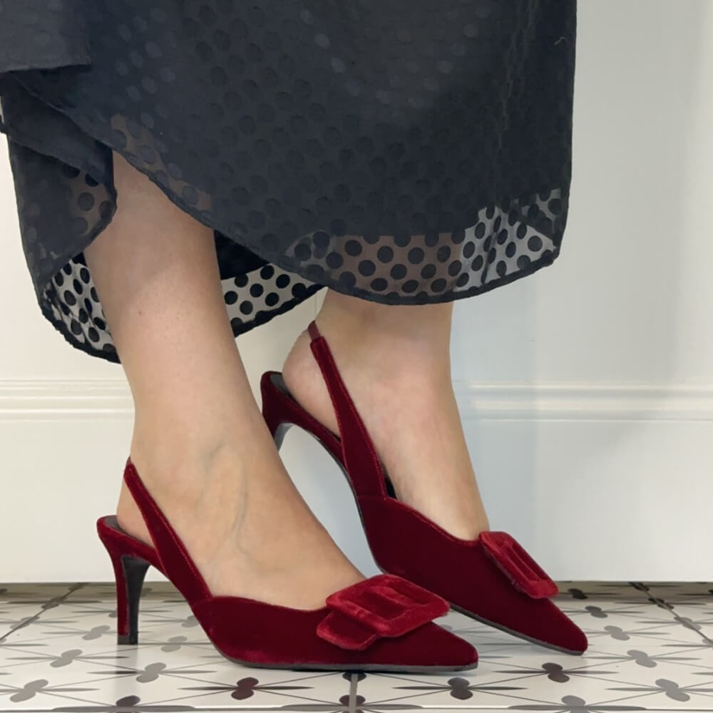 A person wearing Lodi dark red velvet slingback kitten heels with square buckle embellishments on the toes, paired with a sheer black polka dot midi skirt, standing on a patterned tile floor.