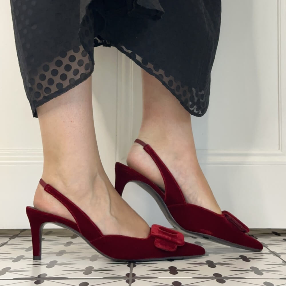 A person wearing Lodi dark red velvet slingback kitten heels with square buckle embellishments on the toes, paired with a sheer black polka dot midi skirt, standing on a patterned tile floor.