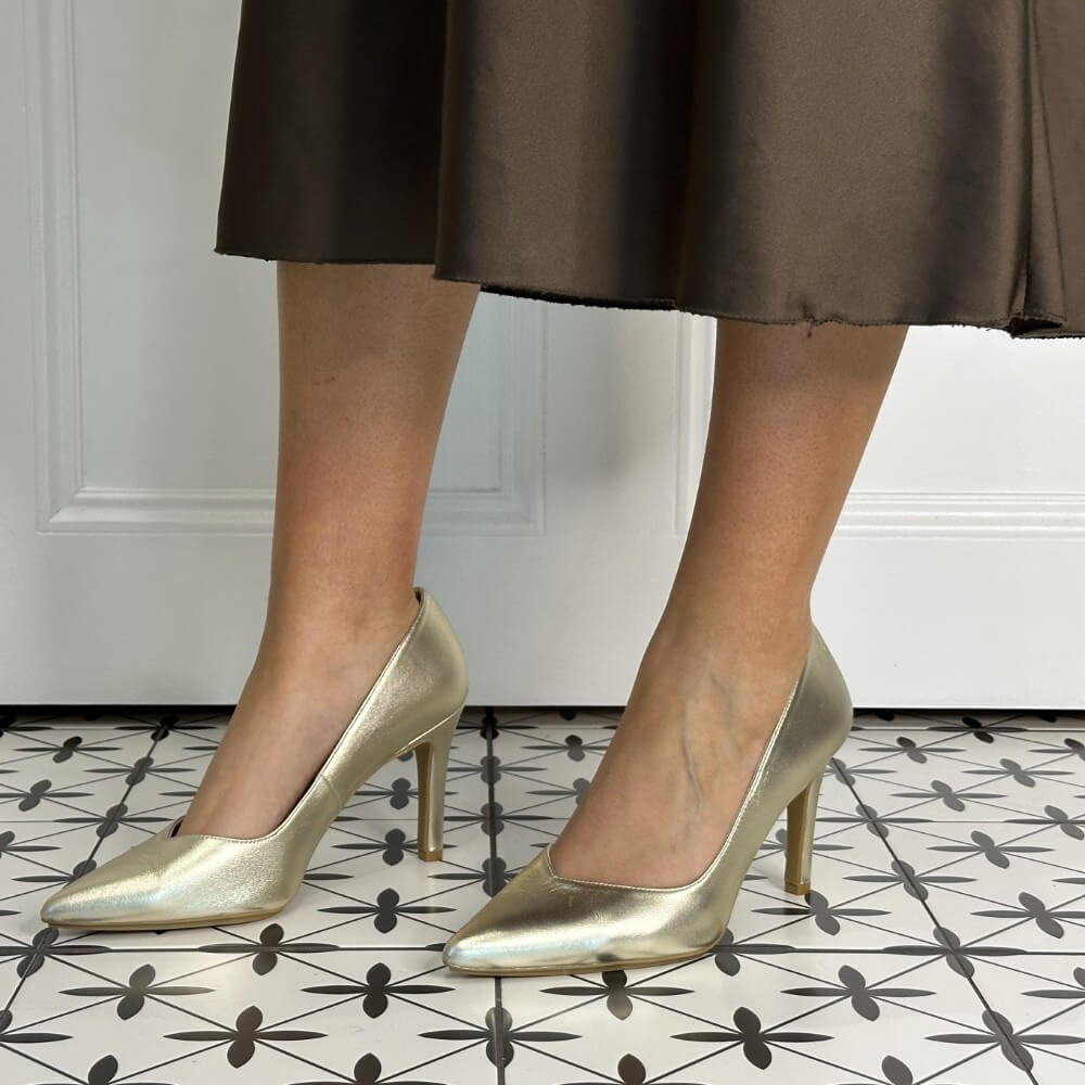Gold high-heeled shoes worn with a brown skirt on a patterned floor.