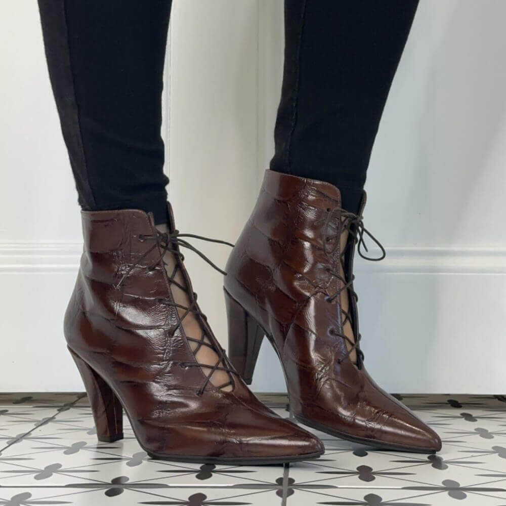 A pair of Lodi dark brown lace-up ankle boots with a crocodile-embossed texture and high stiletto heels, shown on a model wearing black leggings.