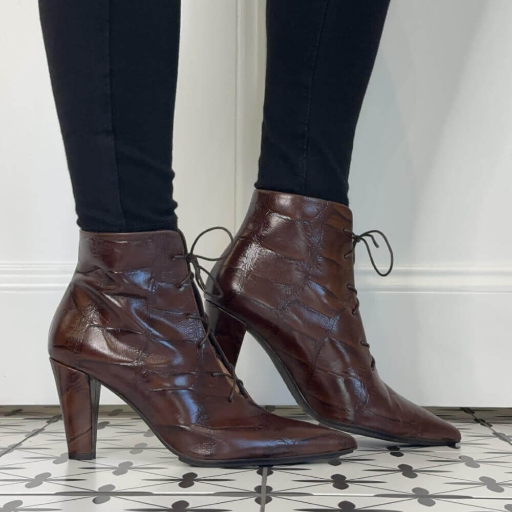 A pair of Lodi dark brown lace-up ankle boots with a crocodile-embossed texture and high stiletto heels, shown on a model wearing black leggings.