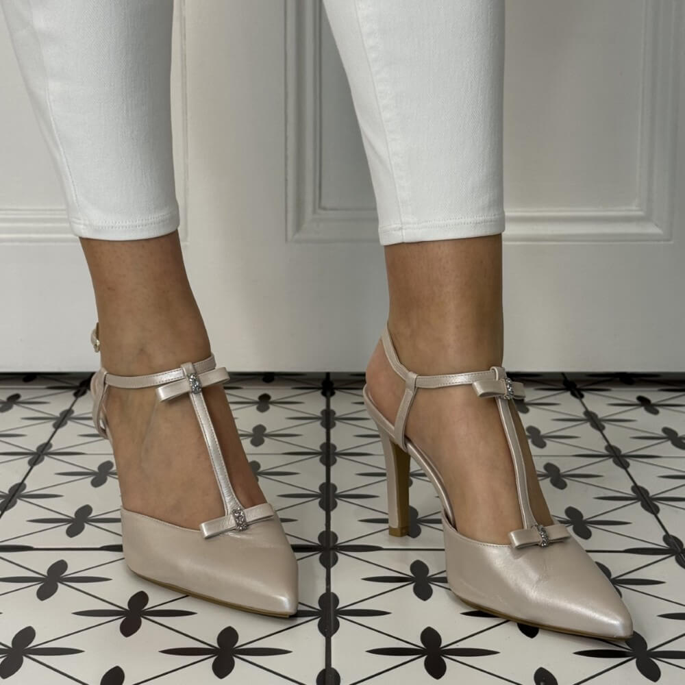 Beige high-heeled shoes with metallic straps worn with white pants on a patterned floor.