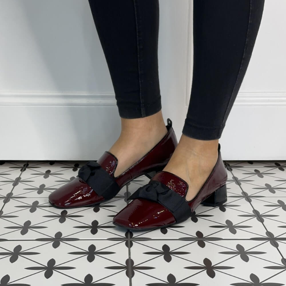 Burgundy shoes with black bows worn with black pants on a patterned floor.
