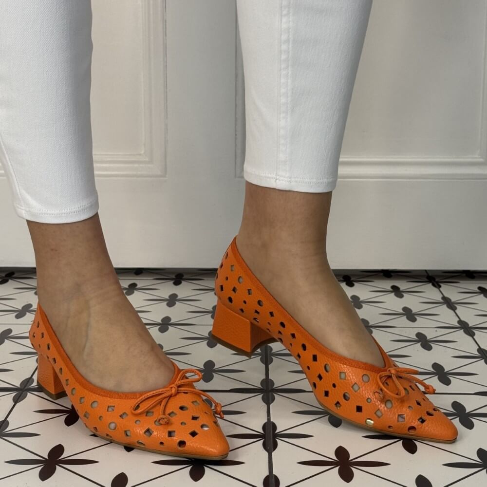 Orange perforated shoes worn with white pants on a patterned floor.
