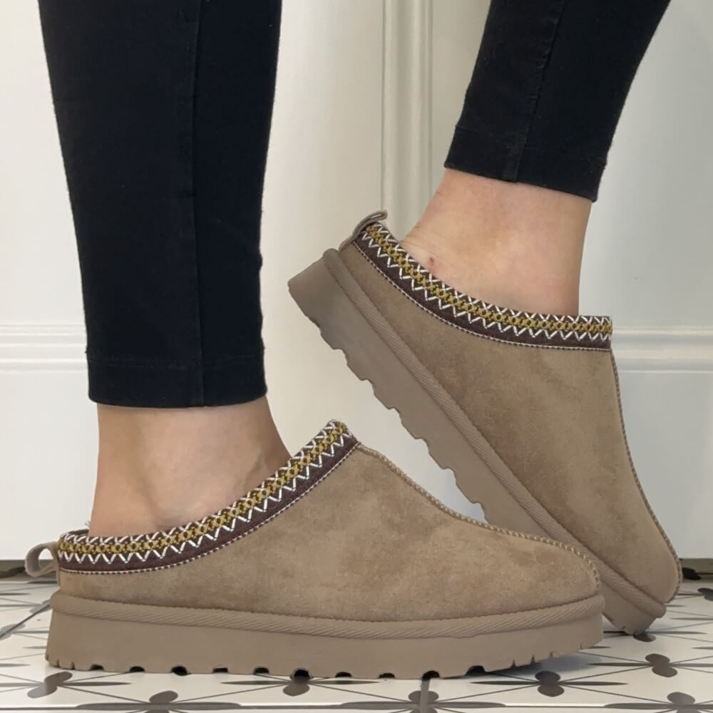 A person wearing black leggings and Lunar taupe suede slip-on slippers with a braided trim detail, standing on a patterned tile floor in front of a white paneled wall.