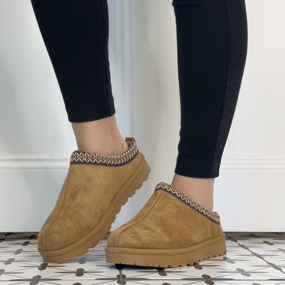 A pair of Lunar tan suede slip-on clog slippers with a patterned fabric trim, shown being worn with black leggings on a patterned tile floor.