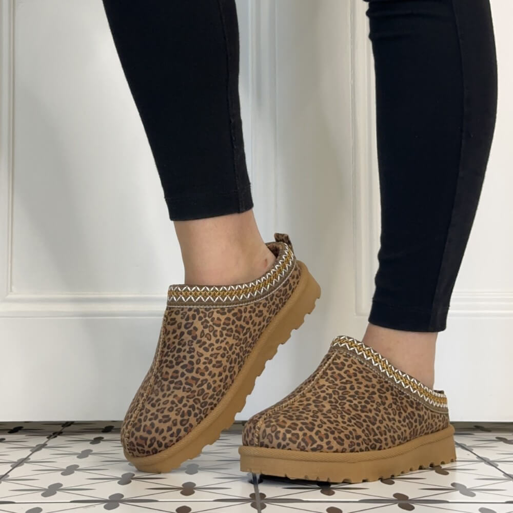 A person wearing Lunar brown leopard-print slip-on clogs with tan soles and a patterned trim, shown from the feet down against a white wall and patterned tile floor.