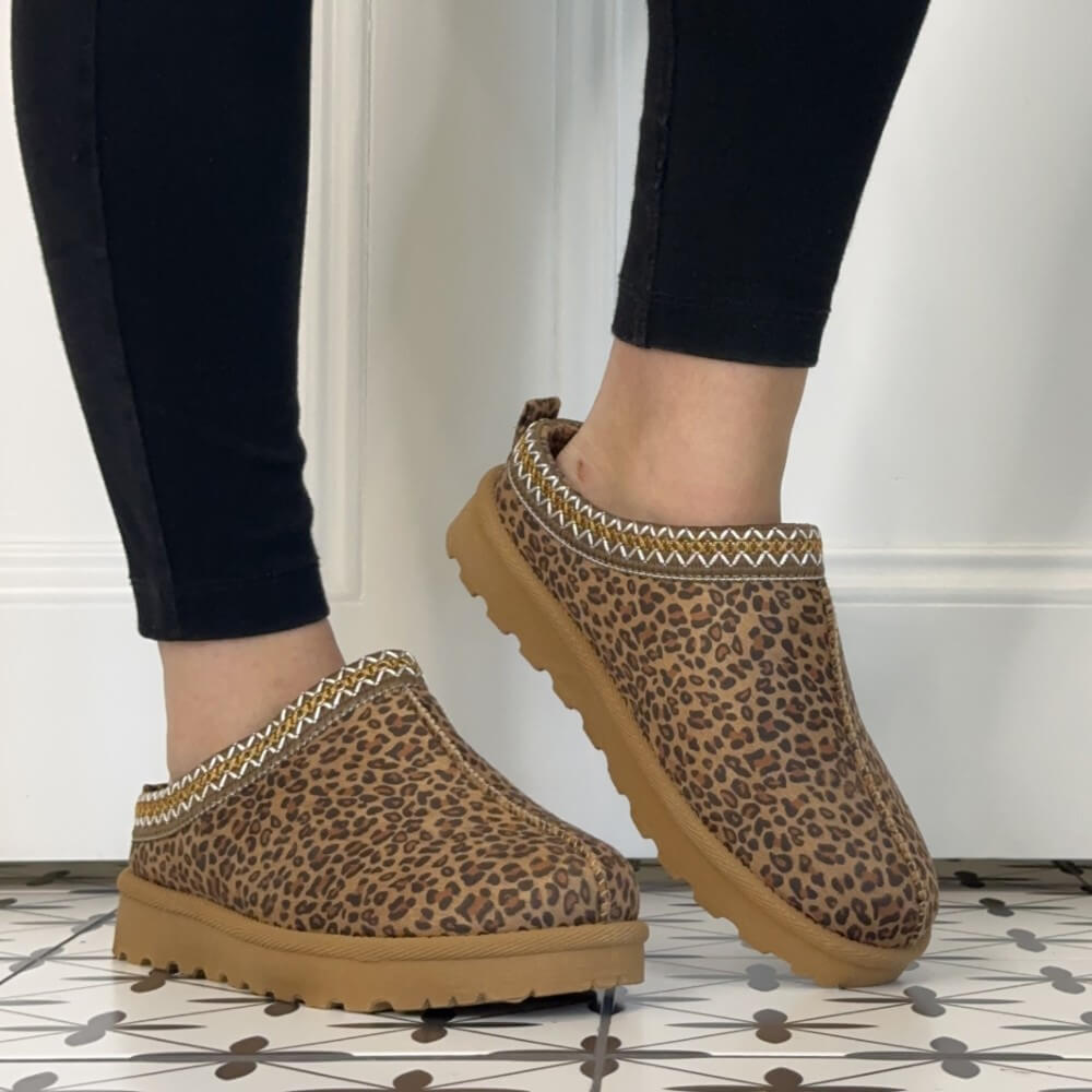 A person wearing Lunar brown leopard-print slip-on clogs with tan soles and a patterned trim, shown from the feet down against a white wall and patterned tile floor.
