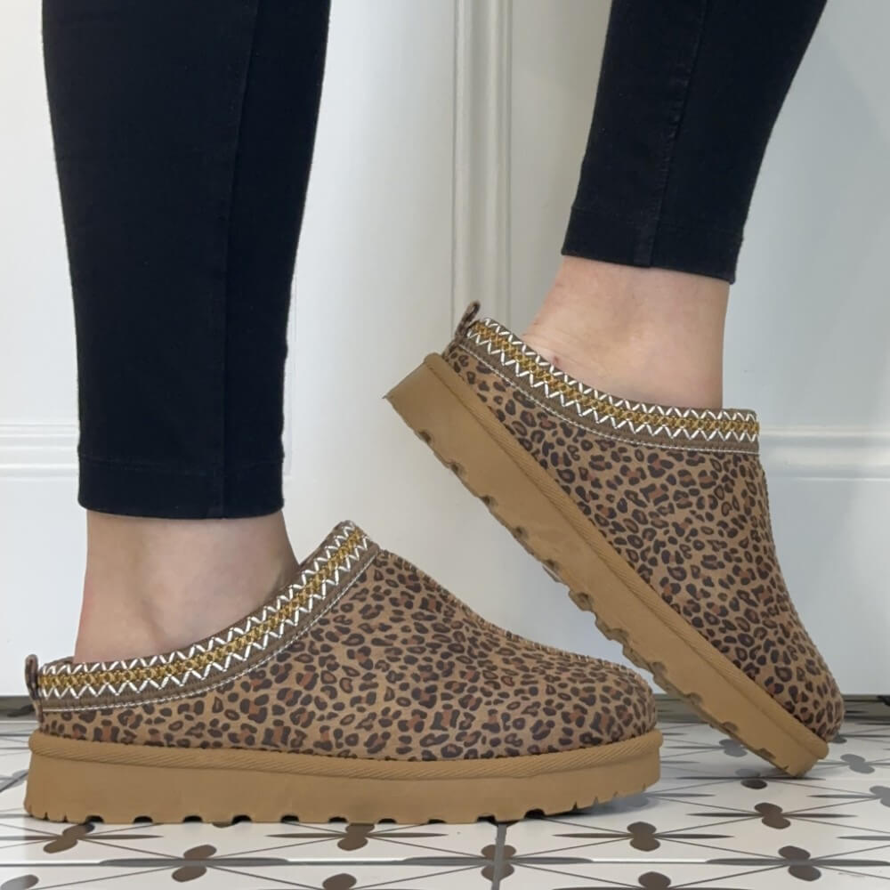 A person wearing Lunar brown leopard-print slip-on clogs with tan soles and a patterned trim, shown from the feet down against a white wall and patterned tile floor.