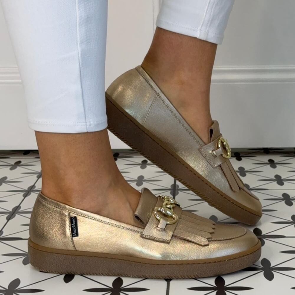 Marco Moreo Gold loafers with fringe details worn by a person on a patterned floor.