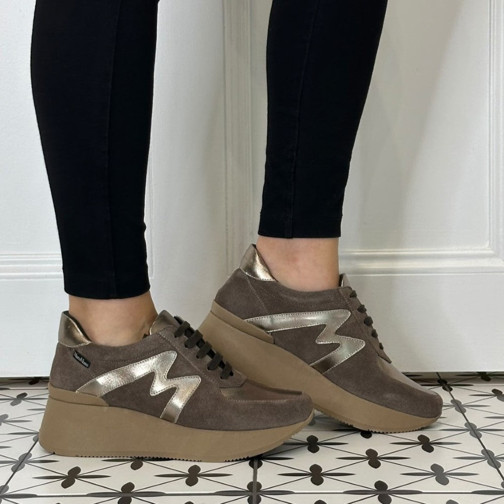 A person wearing Marco Moreo brown suede platform sneakers with metallic silver 'M' logos, paired with black pants, standing on a black and white patterned tile floor against a white panelled wall.